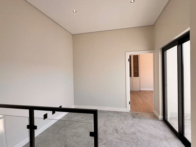 A bright, modern landing or hallway featuring neutral walls, large format floor tiles, and a contemporary glass balustrade with access to an adjacent room.