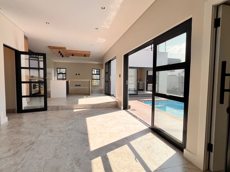 Spacious open-plan living area featuring large black aluminium sliding doors opening to a swimming pool, tiled floors, and a modern timber ceiling detail.