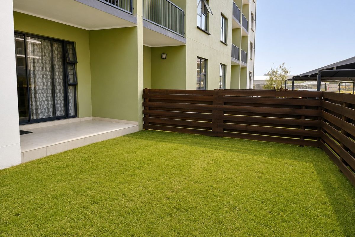 A private ground floor garden area featuring a tiled patio, lawn, and timber slat fencing, adjacent to the apartment building.