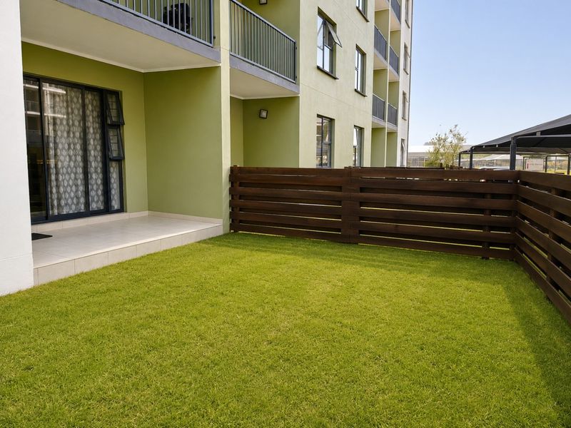 A private ground floor garden area featuring a tiled patio, lawn, and timber slat fencing, adjacent to the apartment building.