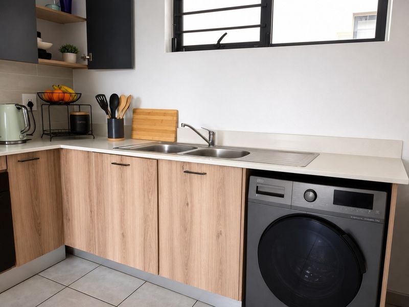 A modern kitchen featuring light wood-finish cabinetry, stone countertops, a double stainless steel sink, and integrated space for a washing machine.