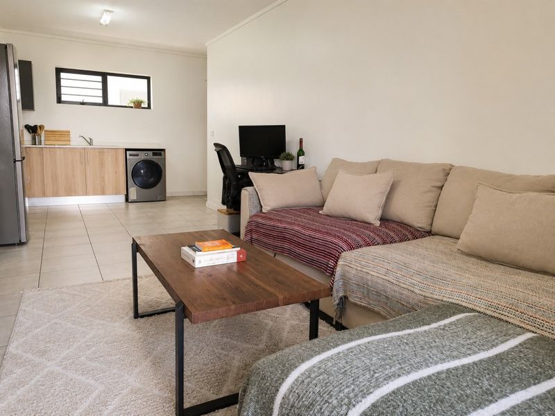 An open-plan living area featuring a functional kitchen corner with cabinetry, sink, and laundry appliance connections, finished with neutral tiled flooring.