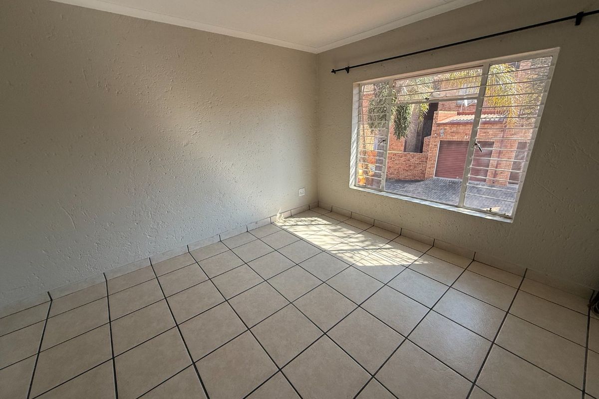 A bright, empty bedroom featuring neutral tiled flooring and a large window fitted with security bars.