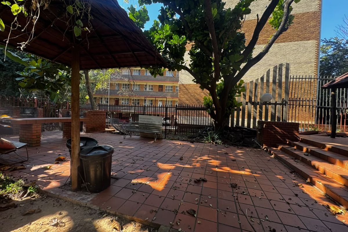 A communal outdoor patio area featuring tiled flooring, a thatched gazebo, and brick seating, enclosed by metal perimeter fencing.
