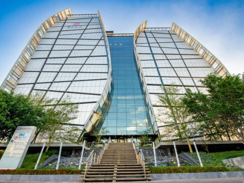 Modern commercial office building featuring a striking glass facade, central atrium, stone staircase entrance, and landscaped water feature.