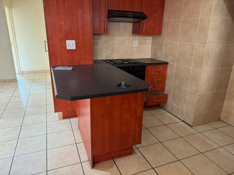 A compact kitchen featuring cherry-wood laminate cabinetry, dark countertops, built-in oven with hob, extractor fan, and neutral ceramic tiling throughout.