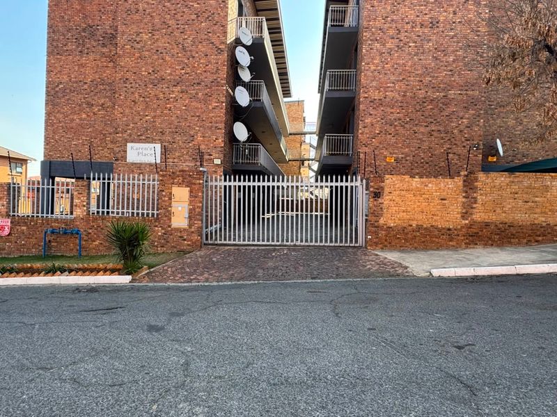 Front exterior view of a brick apartment complex featuring a secure gated entrance, paved driveway, and multiple balconies with satellite dish installations.