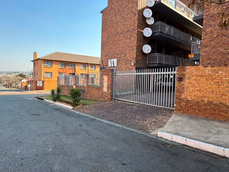 Front exterior view of a multi-story brick apartment building featuring a secure gated entrance, paved driveway, and balcony access.