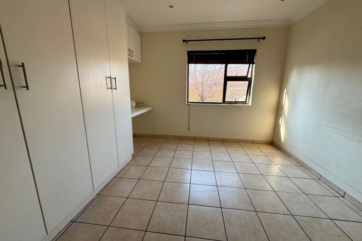 A bright, empty bedroom featuring neutral tiled flooring, built-in cupboards, and a window with a fitted blind.