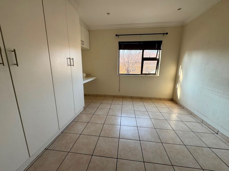 A bright, empty bedroom featuring neutral tiled flooring, built-in cupboards, and a window with a fitted blind.