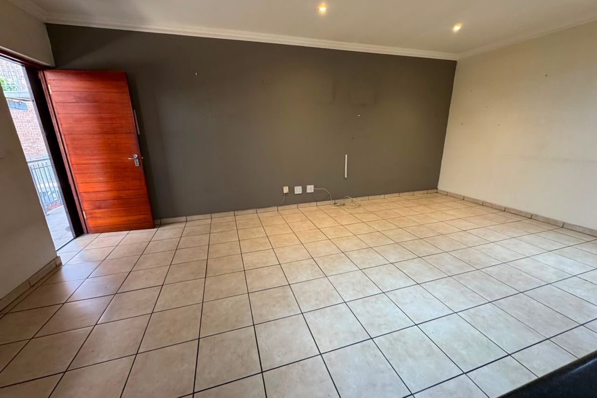 An empty, open-plan living area featuring neutral tiled flooring, recessed lighting, and direct access to an outdoor balcony through a timber door.