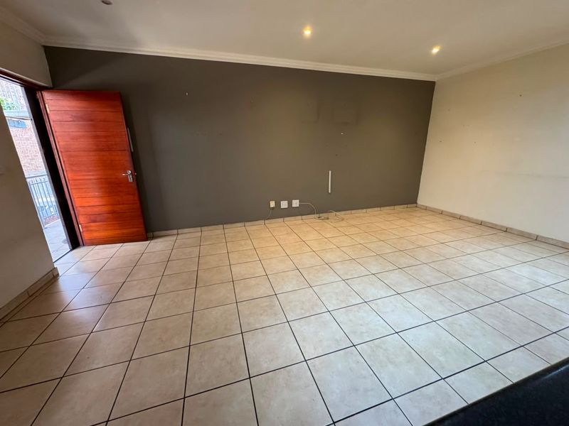 An empty, open-plan living area featuring neutral tiled flooring, recessed lighting, and direct access to an outdoor balcony through a timber door.