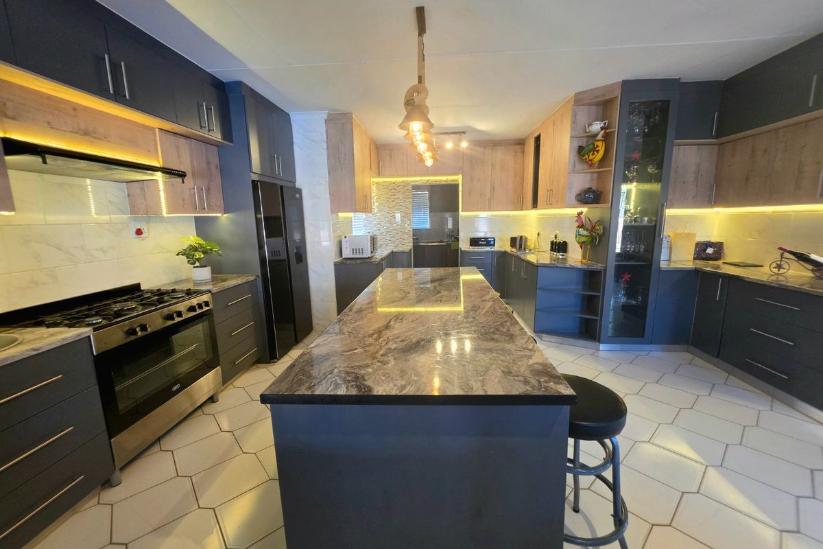 Modern kitchen featuring a large central island, stone countertops, dark cabinetry with wood accents, integrated lighting, and stylish hexagonal floor tiles.