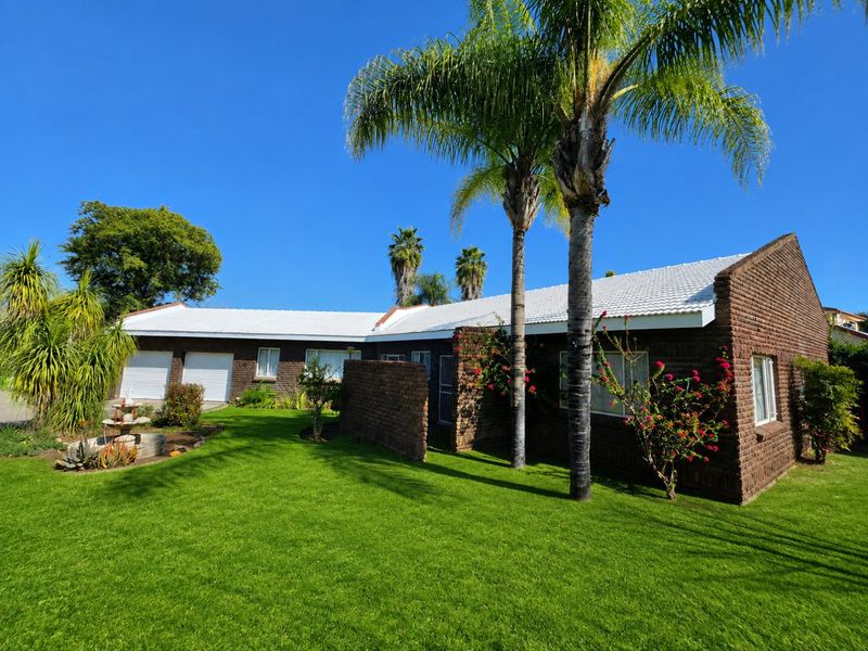 A well-maintained single-story brick house featuring a tiled roof, double garage, and a spacious, manicured front garden with mature palm trees.