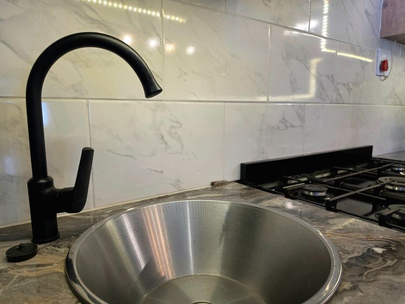 A modern kitchen prep area featuring a stainless steel sink, matte black faucet, gas hob, and marble-look tiled backsplash.