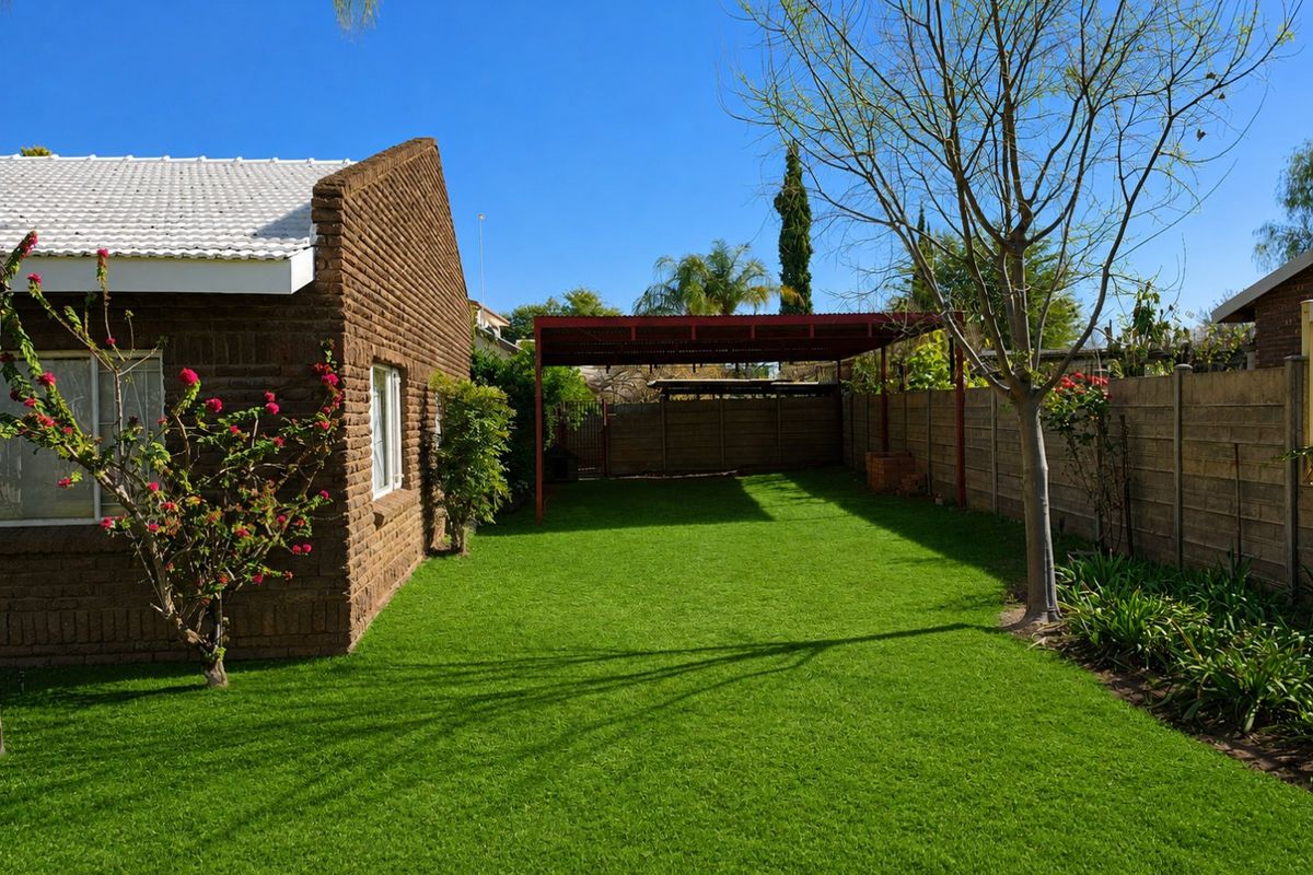 A well-maintained rear garden featuring a neat lawn, brick exterior wall, and a covered metal carport structure.