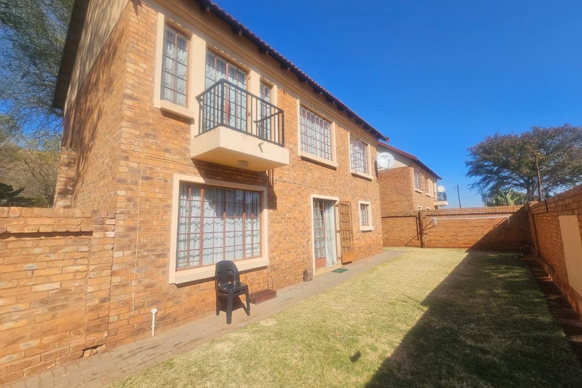 Rear view of a double-story face brick townhouse featuring a small balcony, paved walkway, and a private garden enclosed by brick boundary walls.