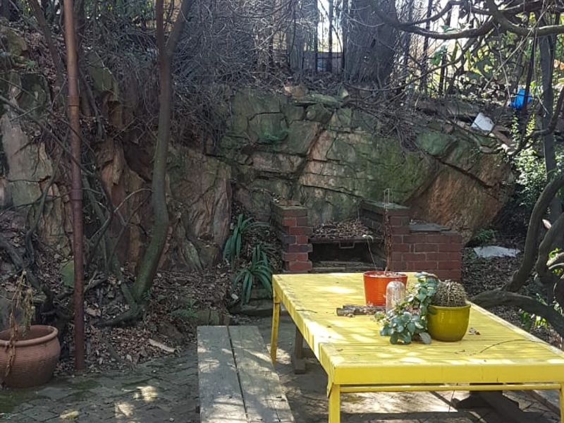 Outdoor patio area featuring brick paving, a built-in brick braai, and a natural rock face backdrop, shaded by an overhead metal pergola structure.
