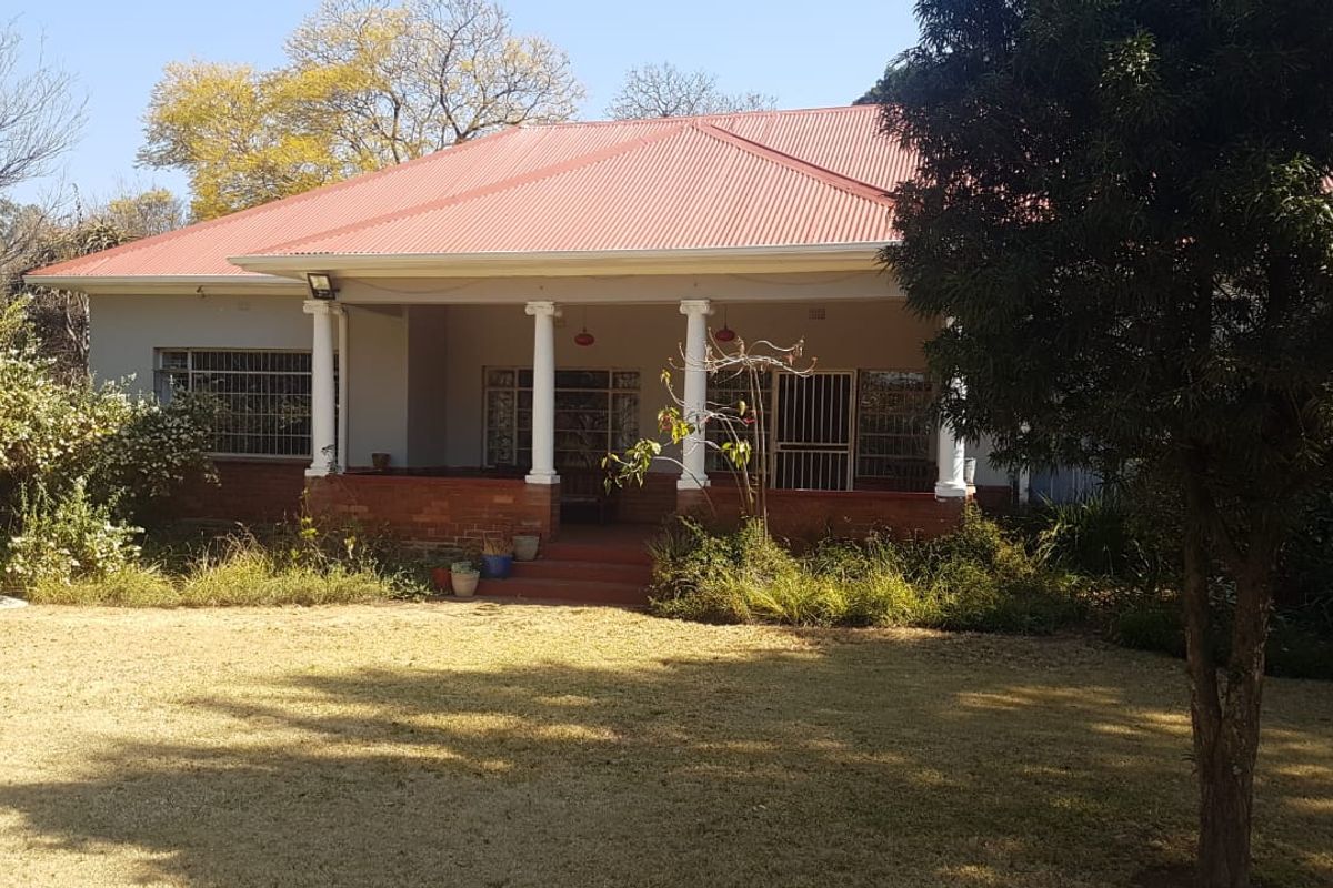 Rear view of a single-story house featuring a covered patio with brick base and columns, set within a large garden area.
