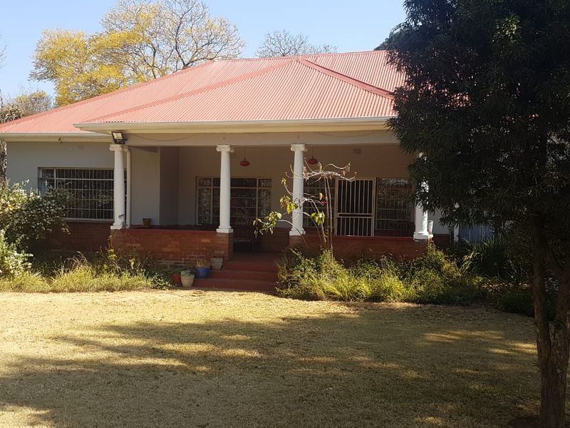 Rear view of a single-story house featuring a covered patio with brick base and columns, set within a large garden area.