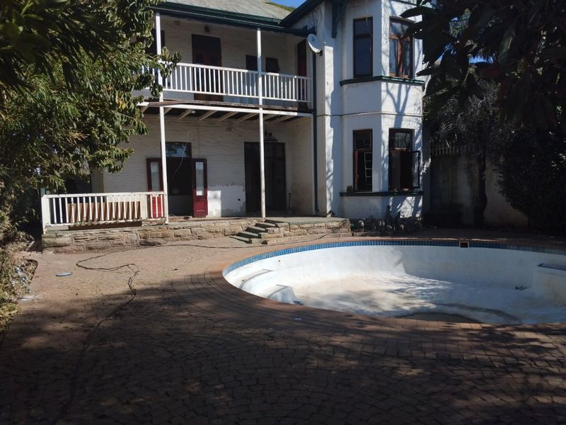 Rear view of a two-story house featuring a balcony, stone retaining wall, and an empty swimming pool set within a paved outdoor area.