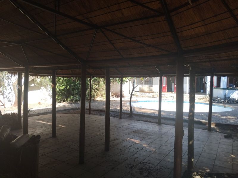 A spacious covered patio area featuring a thatched roof and tiled flooring, overlooking an outdoor swimming pool and garden space.