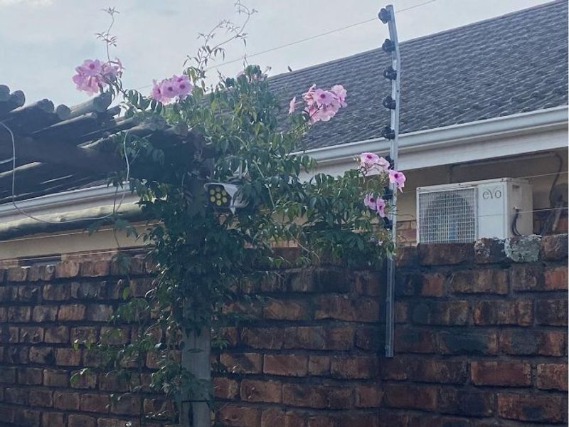 Exterior view of a brick boundary wall featuring integrated electric fencing, a security camera, and an external air conditioning unit.