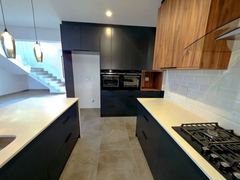 Modern kitchen featuring dark cabinetry, wood-tone accents, stone countertops, a central island, and built-in appliances, with an open-plan layout leading to the staircase.