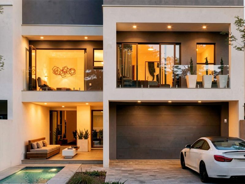 Modern multi-story residence featuring a rear patio, swimming pool, glass balustrades, and an integrated double garage with paved driveway.