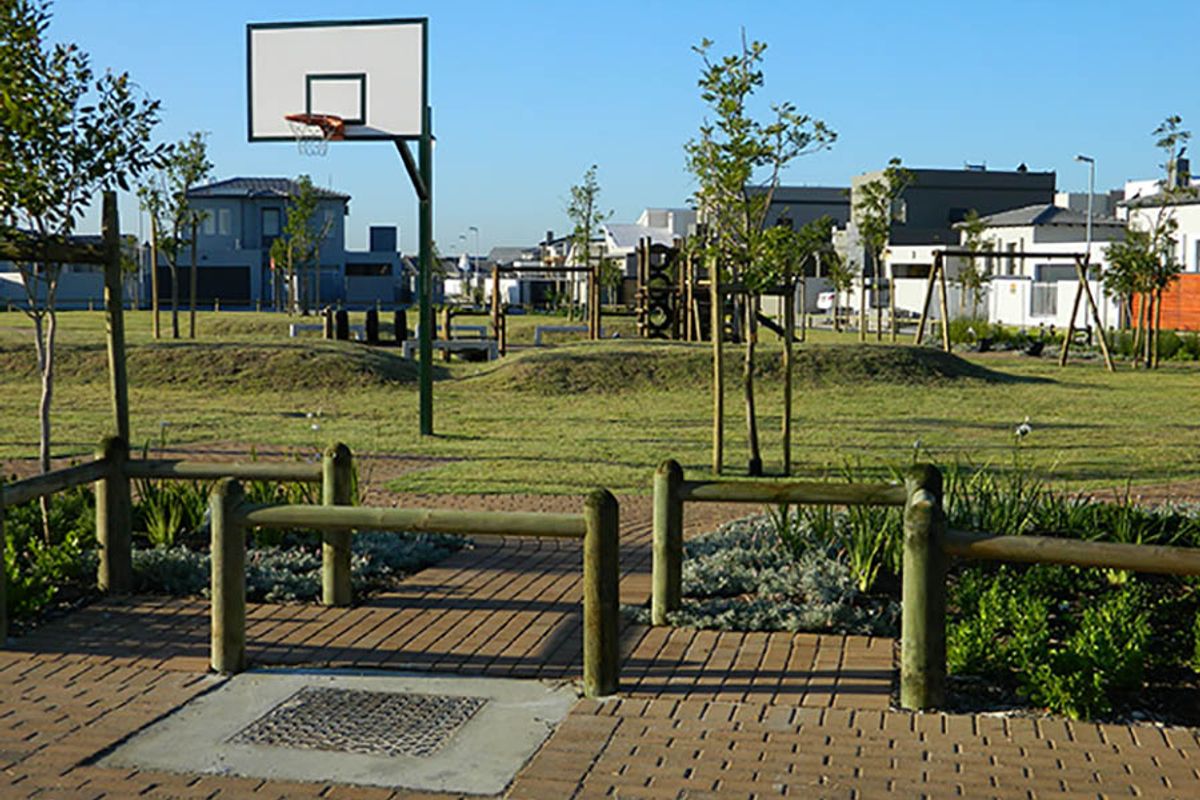 A well-maintained communal park area featuring a basketball hoop, playground equipment, paved pathways, and timber bollard fencing within a residential estate.