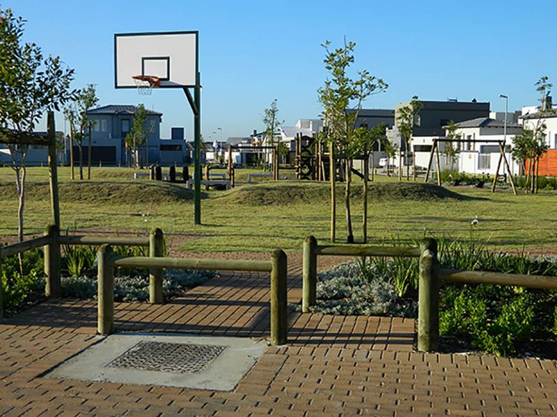 A well-maintained communal park area featuring a basketball hoop, playground equipment, paved pathways, and timber bollard fencing within a residential estate.