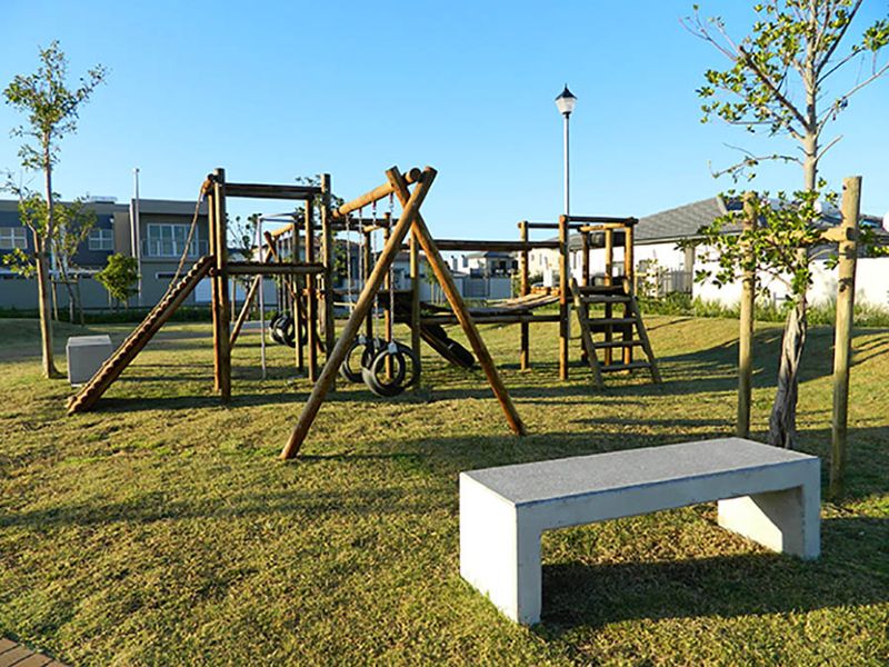 A well-maintained communal garden area featuring a wooden playground structure, a concrete bench, and established landscaping.