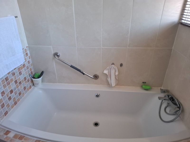 A functional bathroom featuring a built-in bathtub with a chrome grab rail and a combination of neutral and mosaic wall tiling.