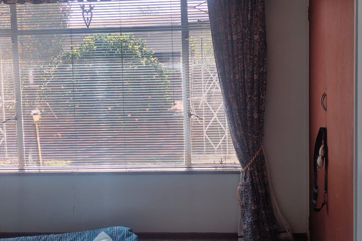 A bedroom featuring a built-in cupboard, large window with venetian blinds, and security bars, offering a view of the garden.