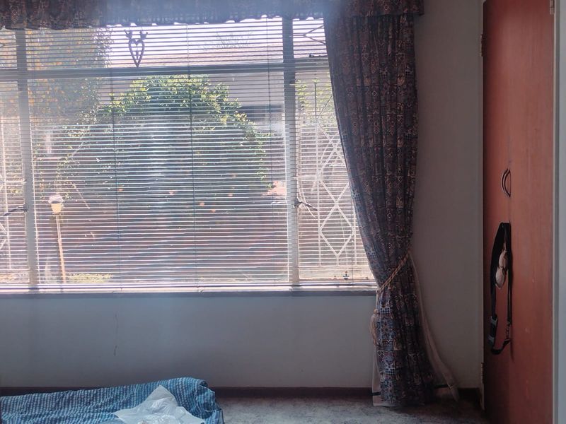 A bedroom featuring a built-in cupboard, large window with venetian blinds, and security bars, offering a view of the garden.