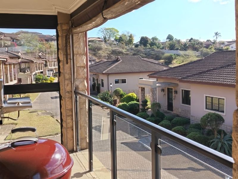 A covered balcony featuring a glass balustrade, stone-clad pillars, and retractable blinds, offering an elevated view of the surrounding townhouse complex.