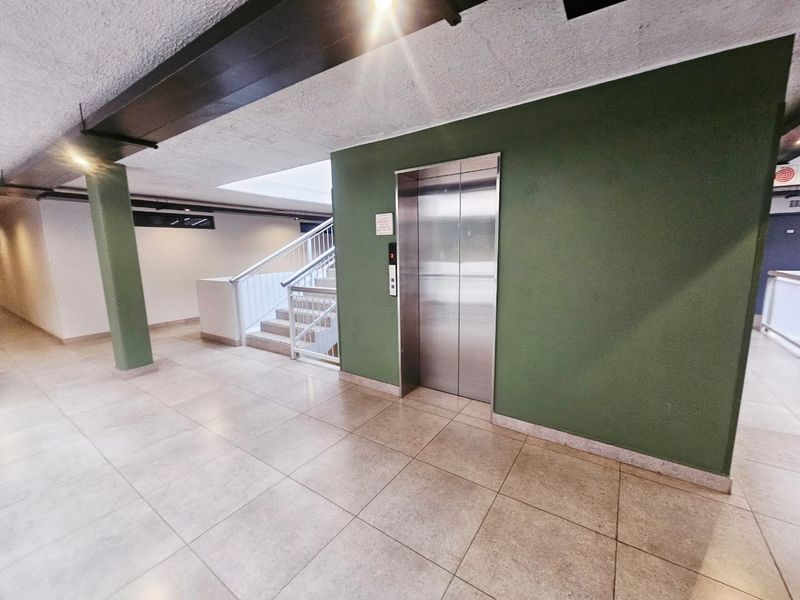 A clean and well-maintained apartment building hallway featuring tiled flooring, a stainless steel elevator, and a staircase with metal handrails.