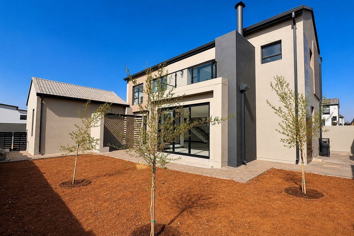 Rear view of a modern two-story house featuring a paved patio, balcony, and landscaped garden area with perimeter security fencing.