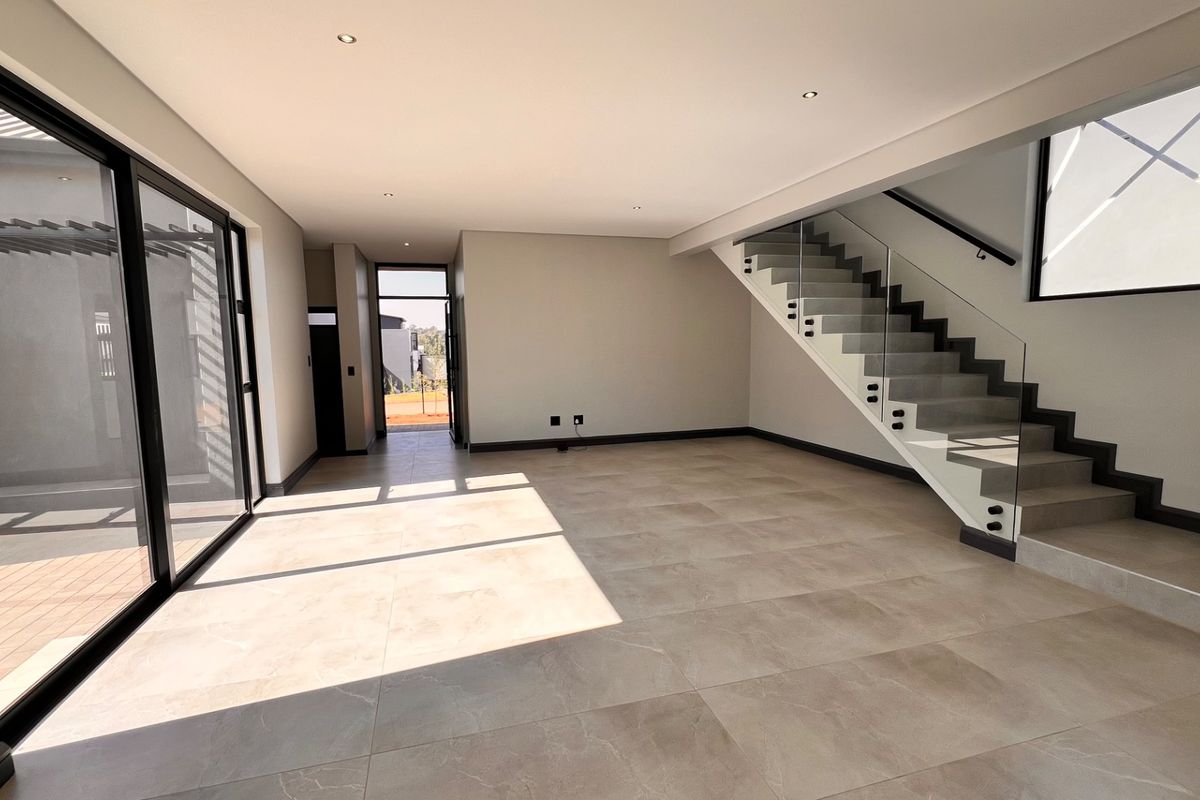 Spacious open-plan living area featuring large format floor tiles, modern staircase with glass balustrade, and sliding doors leading to an outdoor patio.