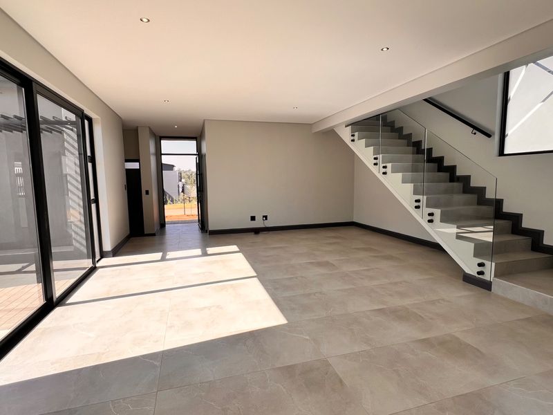 Spacious open-plan living area featuring large format floor tiles, modern staircase with glass balustrade, and sliding doors leading to an outdoor patio.