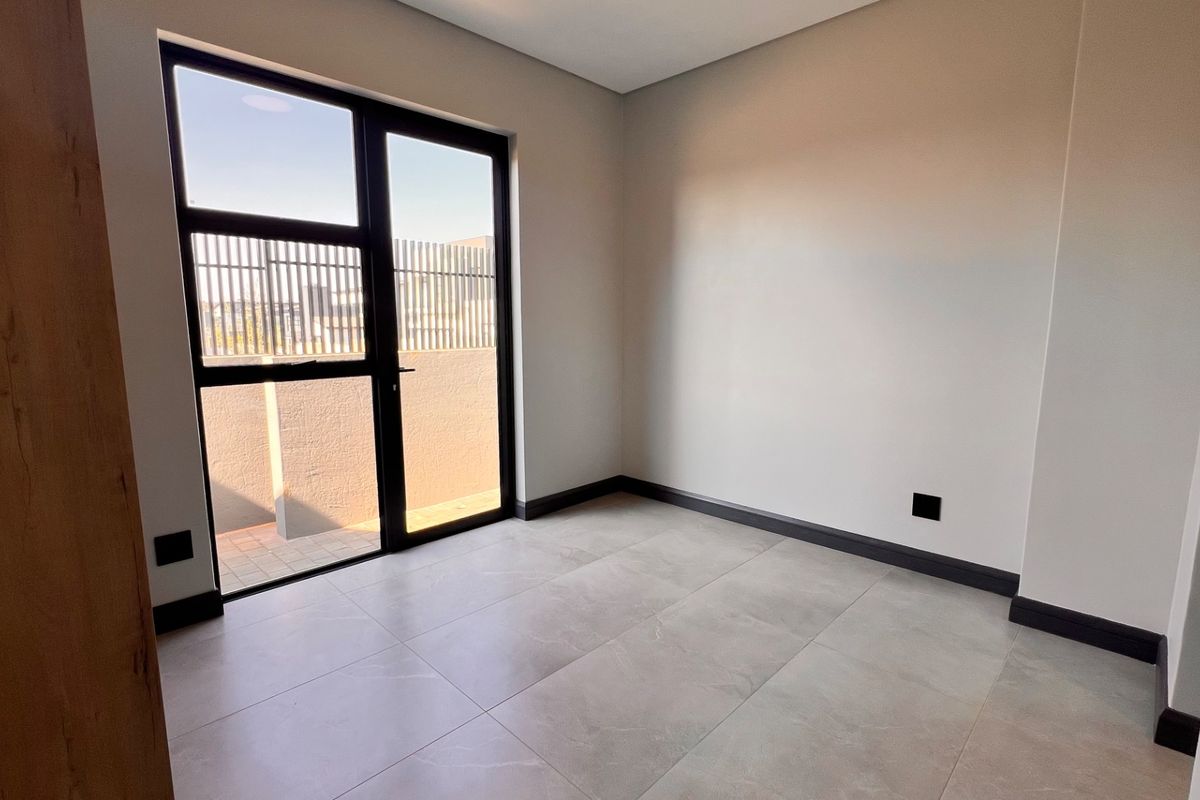 A bright, modern, and empty bedroom featuring large format floor tiles, dark skirting boards, and a glass door providing access to an outdoor area.