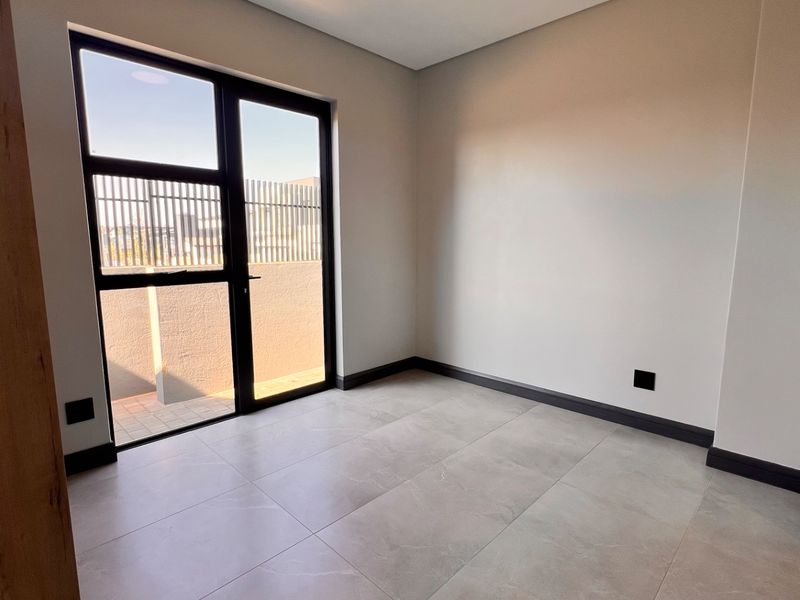 A bright, modern, and empty bedroom featuring large format floor tiles, dark skirting boards, and a glass door providing access to an outdoor area.