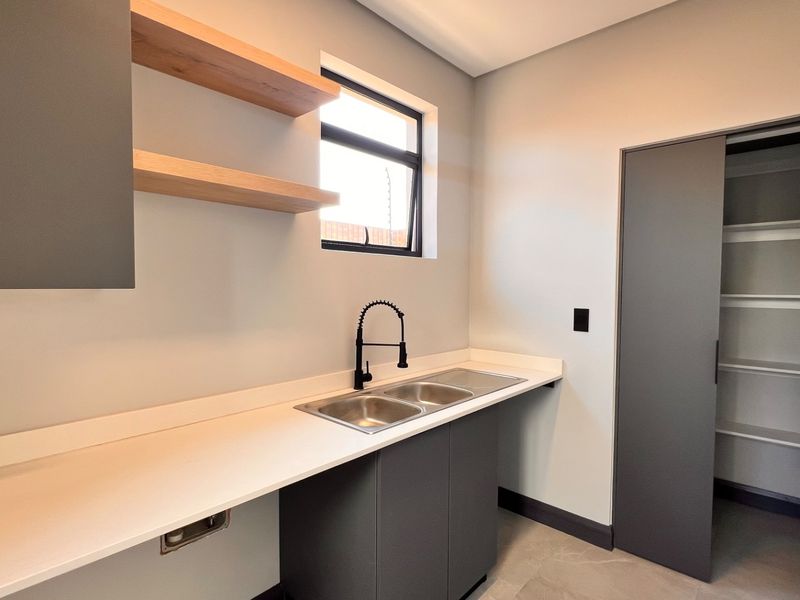 Modern scullery featuring sleek grey cabinetry, stone countertops, a double sink with a gooseneck faucet, floating shelves, and an integrated walk-in pantry.