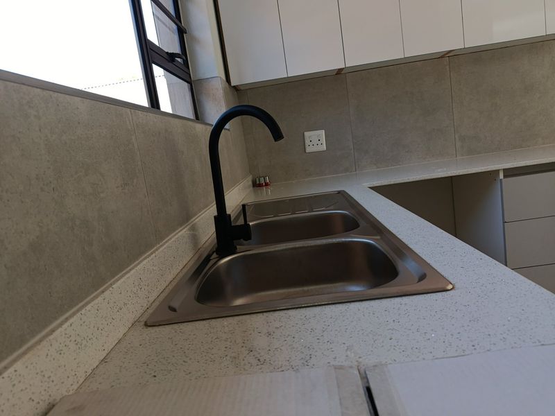 Modern kitchen area featuring a double stainless steel sink, black mixer tap, stone-look countertops, and white handleless cabinetry with grey tiled splashbacks.