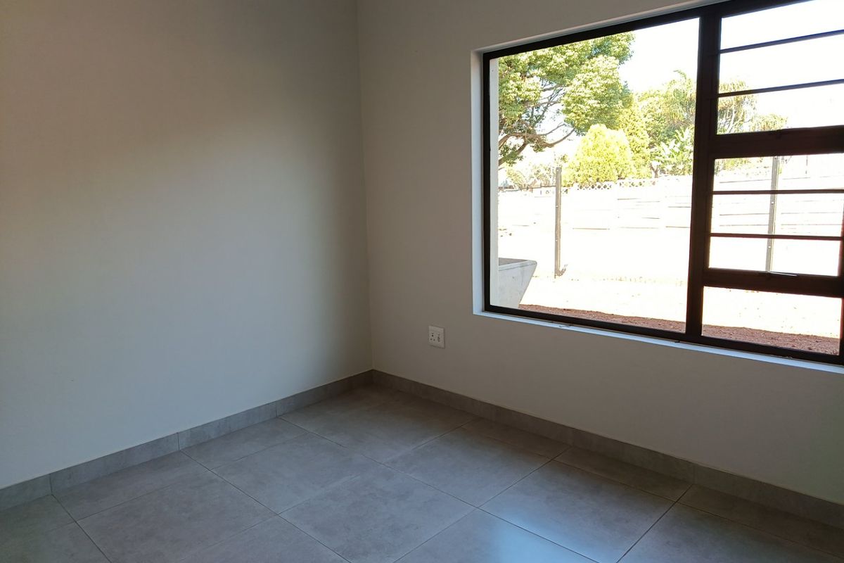 A bright, empty bedroom featuring neutral tiled flooring, clean walls, and a large window with security bars overlooking the garden.