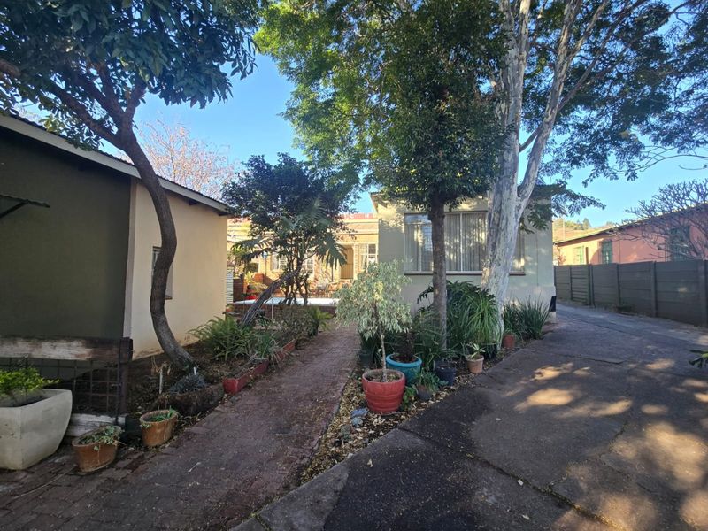 Rear exterior view of the property featuring a brick paved pathway, concrete driveway, and established garden area with mature trees.
