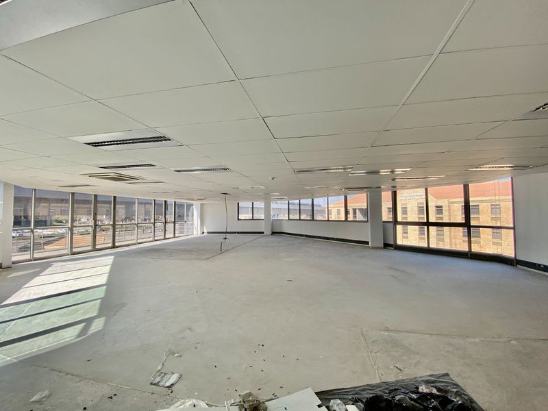 Spacious open-plan commercial office area featuring large floor-to-ceiling windows, suspended ceilings with recessed lighting, and unfinished concrete flooring.