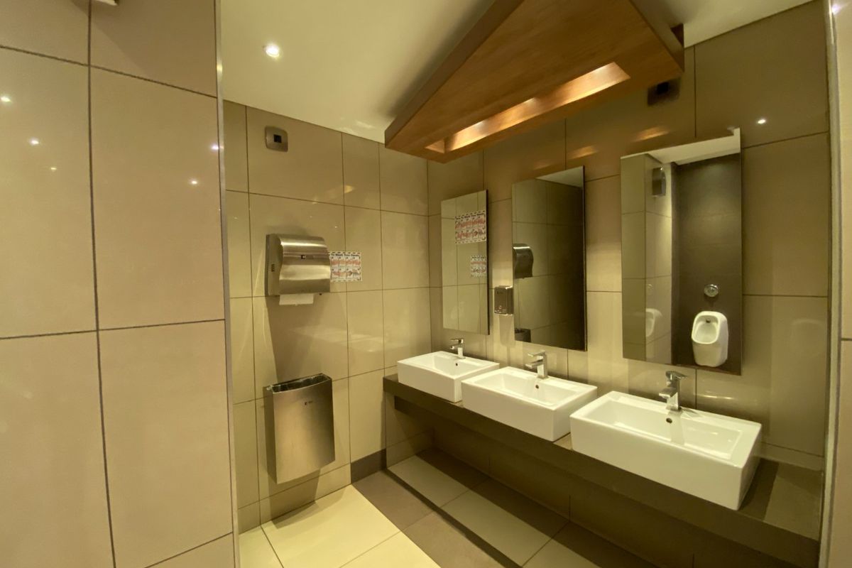 Modern commercial bathroom featuring a triple vanity setup with wall-mounted mirrors, tiled finishes, and integrated stainless steel fixtures.