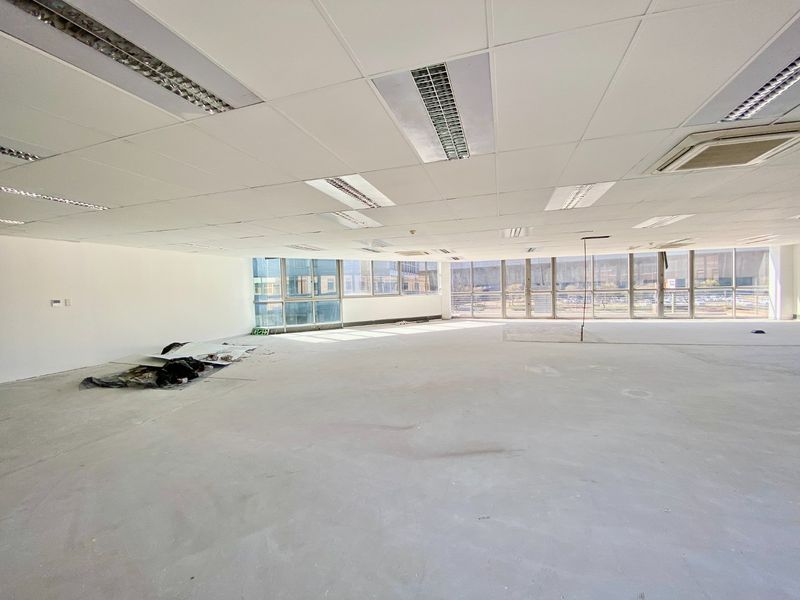 A large, open-plan commercial office space featuring a suspended ceiling, recessed lighting, and expansive floor-to-ceiling windows with views of the surrounding area.