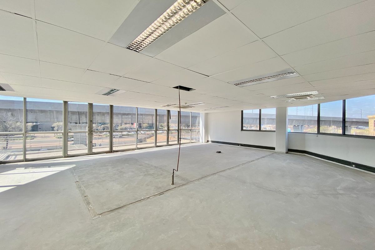 A vacant, open-plan commercial office space featuring floor-to-ceiling windows, suspended ceilings with recessed lighting, and unfinished concrete flooring.